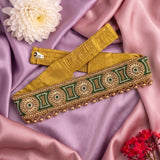 A mustard yellow embroidered waist belt with green and gold geometric patterns and bead trim, laid on draped pink satin fabric with decorative flowers.