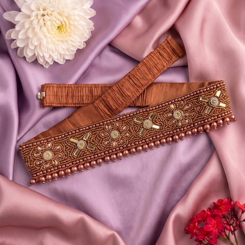 An ornate brown beaded belt with gold and green embellishments, laid out on draped pink satin fabric with decorative flowers.