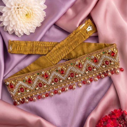 An ornate gold embroidered waist belt or sash laid flat on draped lavender satin fabric, decorated with red and white beadwork, rhinestones, and dangling red and pearl-like beads. A white flower and small red flowers are placed as styling props.