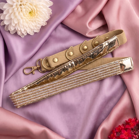 Two decorative straps laid on draped satin fabric: one tan canvas strap with gold-tone hardware and one metallic gold strap covered in small clear rhinestones. White and red flowers are placed in the corners as styling props.