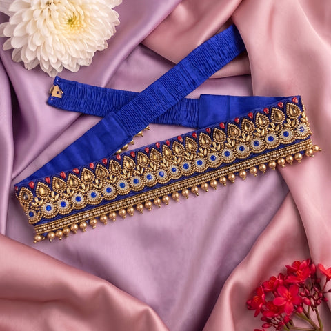 A royal blue waist belt with ornate gold embroidery, blue and red accents, and gold bead borders, displayed on draped pink satin fabric with decorative flowers.