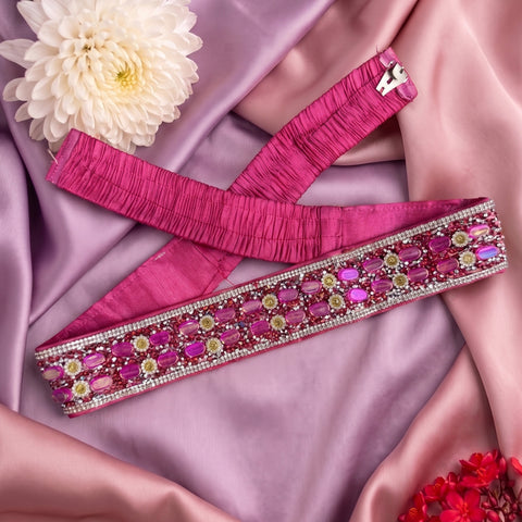 A bright pink embellished sash or belt laid diagonally on draped pink satin fabric, with a white flower and small red flowers as styling props.
