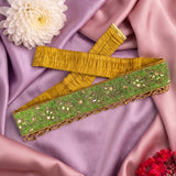 A wide decorative fabric trim or sash laid flat on draped lavender satin, with a yellow-gold pleated inner side and a green outer panel decorated with sequins, beads, and metallic embroidery. White and red flowers are placed around it as styling props.