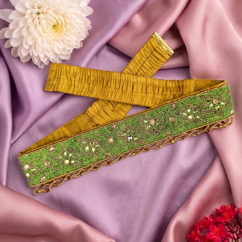 A wide decorative fabric trim or sash laid flat on draped lavender satin, with a yellow-gold pleated inner side and a green outer panel decorated with sequins, beads, and metallic embroidery. White and red flowers are placed around it as styling props.