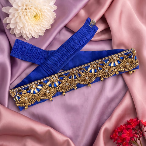 A royal blue fabric waist belt or sash with an ornate gold beaded and embroidered border, laid flat on draped pink satin fabric with decorative flowers.