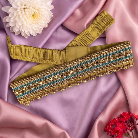 An embroidered fabric trim or sash laid flat on draped lavender satin, with a white flower and small red flowers as styling props. The trim is olive-gold with turquoise blue stripes and metallic gold beadwork and sequins.