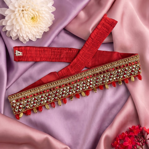 A red and gold embellished waist belt with red and gold tassels, laid flat on draped pink and lavender satin fabric with decorative flowers.