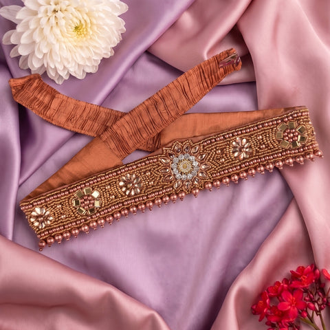 An embroidered peach-brown waistband or belt laid flat on draped pink satin fabric, decorated with metallic gold beadwork, floral motifs, and small pearl-like beads, with white and red flowers as styling props.