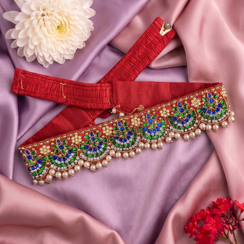 A red fabric waist belt or sash with ornate gold, green, and blue embroidered floral patterns and pearl bead trim, arranged on pink satin fabric with decorative flowers.