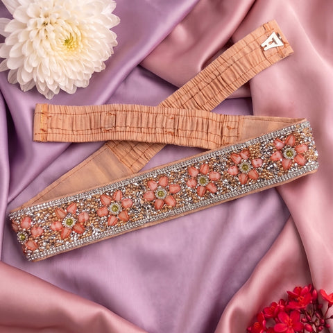 A peach-beige embellished sash laid diagonally on draped pink satin fabric, with a white flower and small red flowers as styling props. The sash has floral beadwork in pink, peach, and green with rhinestone trim and elastic ends.