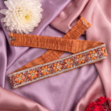 An orange-brown elastic waistband with a wide, heavily embellished decorative band, laid flat on draped pink satin fabric with a white flower prop.