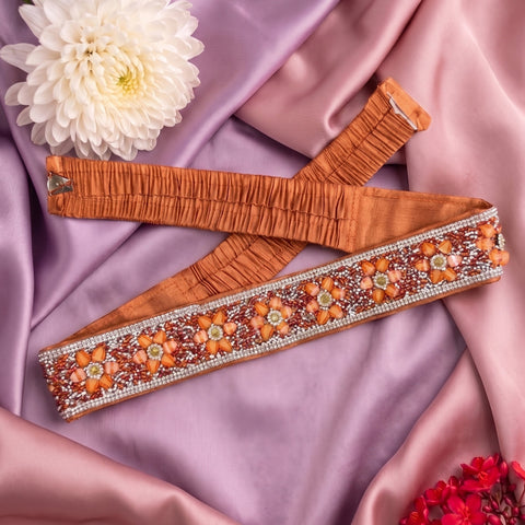 An orange-brown elastic waistband with a wide, heavily embellished decorative band, laid flat on draped pink satin fabric with a white flower prop.