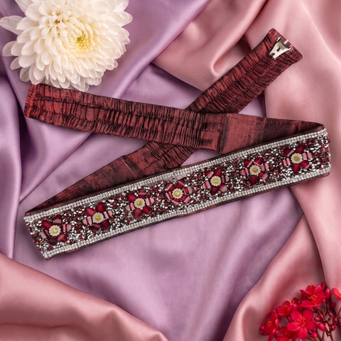 A wide waist belt with a textured burgundy fabric body and an ornate beaded floral border, laid out on draped pink satin fabric with decorative flowers.