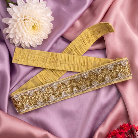 A mustard yellow fabric belt with intricate gold and silver beaded embroidery, laid out on a draped pink satin fabric with decorative white and red flowers.