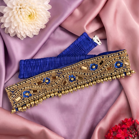 A royal blue pleated waist belt with ornate gold beaded embroidery and blue floral motifs, laid flat on a draped pink satin fabric with decorative flowers.