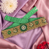 A green and gold embroidered traditional waist belt or decorative sash laid flat on pink satin fabric, with a white flower and small red flowers as styling props.