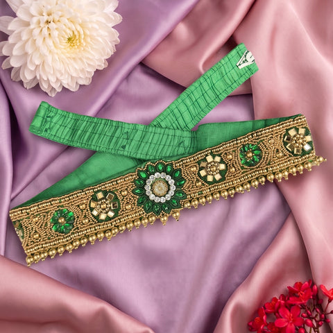 A green and gold embroidered traditional waist belt or decorative sash laid flat on pink satin fabric, with a white flower and small red flowers as styling props.
