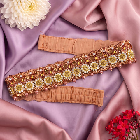 A decorative embroidered waistband or belt laid flat on draped pink satin fabric, with floral and bead embellishments in gold, white, and pink. Two matching fabric bands are visible, styled with flowers in the corners.