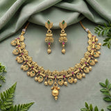 An ornate gold-toned traditional necklace and matching drop earrings laid flat on a soft sage green fabric background, accented with small green and pink stones and surrounded by green leaves.