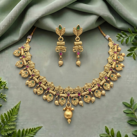An ornate gold-toned traditional necklace and matching drop earrings laid flat on a soft sage green fabric background, accented with small green and pink stones and surrounded by green leaves.