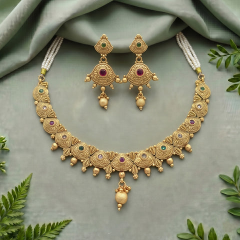 Gold-toned traditional Indian jewelry set featuring a necklace and matching earrings with floral motifs, small green and purple stones, and white pearl-like strands, arranged on a sage green fabric background with decorative leaves.