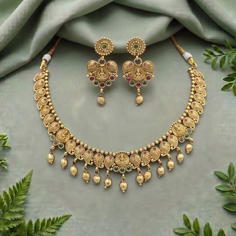 Gold-toned traditional Indian jewelry set featuring a necklace and matching earrings with intricate filigree, small green and pink stones, and dangling bead elements, arranged on a draped sage green fabric with decorative green leaves.