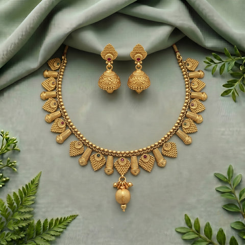 Gold-toned traditional necklace and matching jhumka earrings arranged on a soft sage green fabric backdrop with decorative green leaves around the edges.