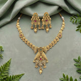 Gold-toned traditional Indian jewelry set featuring a necklace and matching earrings with intricate filigree work, red gemstone accents, and white bead drops, arranged on a sage green draped fabric with decorative green leaves.