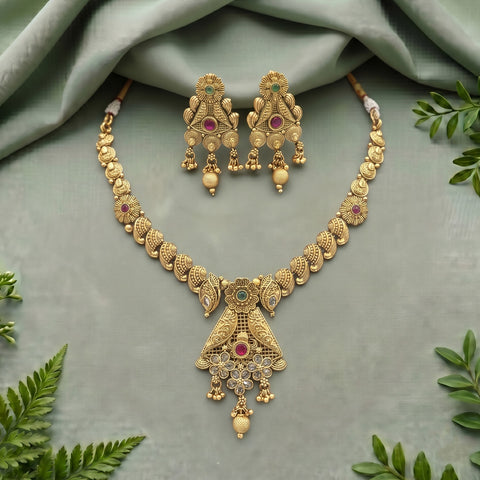 Gold-toned traditional Indian jewelry set featuring a necklace and matching earrings with intricate filigree work, red gemstone accents, and white bead drops, arranged on a sage green draped fabric with decorative green leaves.