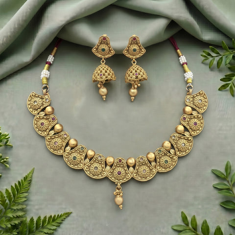 Gold-toned traditional jewelry set featuring a necklace and matching earrings with ornate filigree details, small green and red stone accents, and beaded cords, arranged on a draped sage green fabric with decorative leaves.