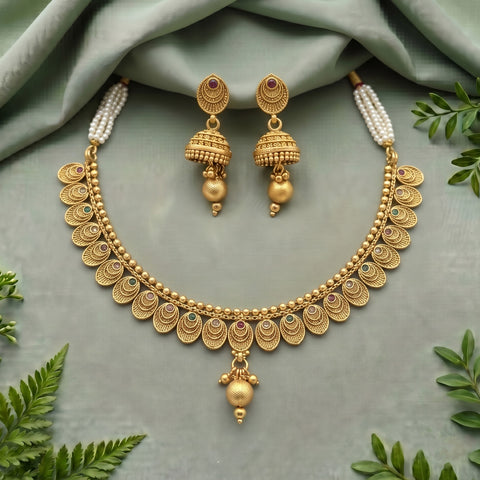 Gold-toned traditional jewelry set with a necklace and matching earrings, arranged on a soft sage green fabric backdrop with decorative green leaves around the edges. The necklace has a scalloped pendant border, beaded details, pearl-like side strands, and a dangling ornament at the center, while the earrings mirror the pendant design.