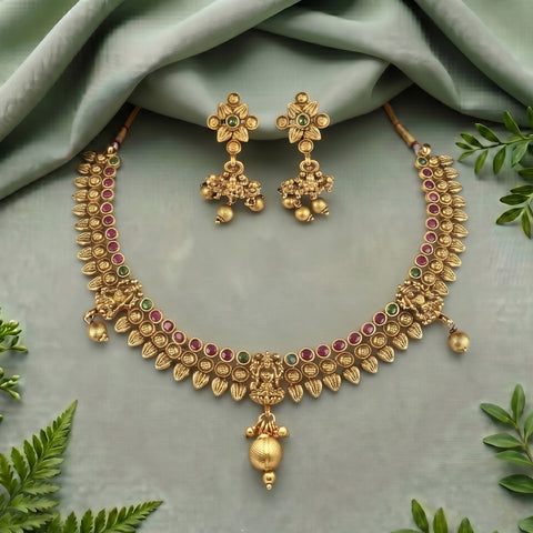 Gold-toned traditional Indian jewelry set featuring a necklace and matching earrings, embellished with red and green stones, displayed on a sage green fabric background with decorative green leaves.