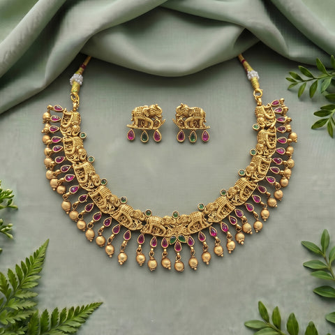 Gold-toned traditional necklace and matching earrings with elephant motifs, pink gemstone teardrops, and small green stone accents, arranged on a soft sage-green fabric backdrop with decorative leaves around the edges.