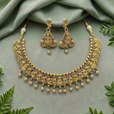 An ornate gold-toned traditional necklace and matching earrings set with small multicolored gemstone accents, arranged on a soft sage green fabric backdrop with decorative green leaves around the edges.