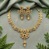 Gold-toned traditional jewelry set with a necklace and matching earrings, decorated with red and green stones and small clear stones, arranged on a soft sage green fabric with decorative green leaves around the edges.