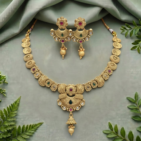 Gold-toned traditional jewelry set with a necklace and matching earrings, decorated with red and green stones and small clear stones, arranged on a soft sage green fabric with decorative green leaves around the edges.