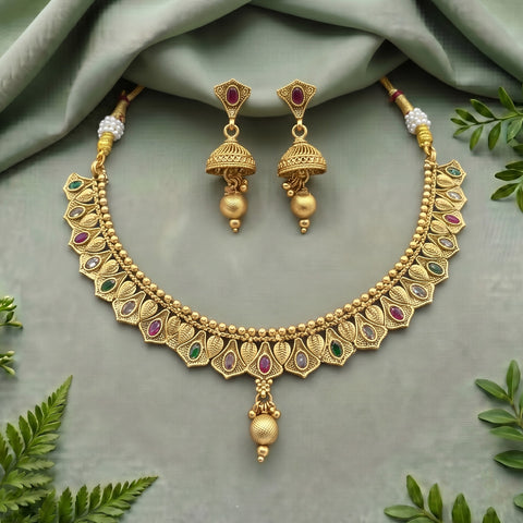 Gold-toned traditional Indian jewelry set featuring a necklace and matching earrings, embellished with red and green stones, displayed on a sage green fabric background with decorative green leaves.