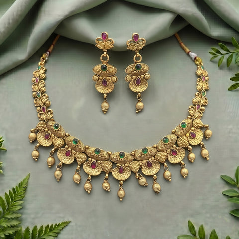 Gold-toned traditional jewelry set featuring a necklace and matching earrings with floral motifs, green stone accents, and small red stone details, arranged on a soft sage green fabric background with decorative leaves.