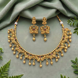 An ornate gold-toned traditional necklace and matching drop earrings displayed on a soft sage green fabric backdrop with decorative green leaves around the edges. The jewelry features detailed filigree, beaded chains, dangling bead drops, and small red and green stone accents.