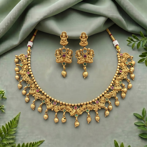 An ornate gold-toned traditional necklace and matching drop earrings displayed on a soft sage green fabric backdrop with decorative green leaves around the edges. The jewelry features detailed filigree, beaded chains, dangling bead drops, and small red and green stone accents.