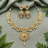 Gold-toned traditional jewelry set featuring a floral choker necklace and matching jhumka earrings, decorated with small red and green stones, displayed on a soft sage-green fabric background with decorative green leaves.