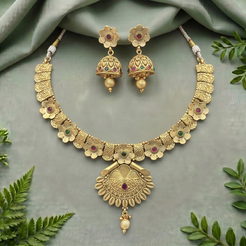 Gold-toned traditional jewelry set featuring a floral choker necklace and matching jhumka earrings, decorated with small red and green stones, displayed on a soft sage-green fabric background with decorative green leaves.