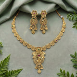 Gold-toned traditional jewelry set featuring a necklace and matching earrings with floral motifs and small green and pink stone accents, arranged on a soft sage-green fabric background with decorative green leaves around the edges.