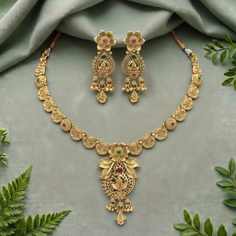 Gold-toned traditional jewelry set featuring a necklace and matching earrings with floral motifs and small green and pink stone accents, arranged on a soft sage-green fabric background with decorative green leaves around the edges.