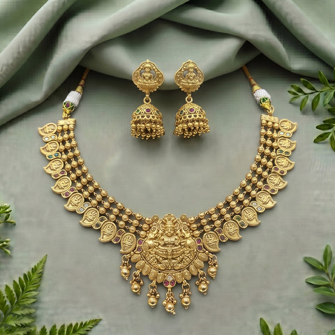 Gold-toned traditional Indian jewelry set featuring a statement necklace and matching dangle earrings, styled on a sage green draped fabric background with decorative green leaves.