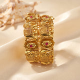 A pair of gold-toned bangle bracelets with detailed floral and geometric engravings and small red gemstone accents, displayed on draped cream satin fabric with a seashell prop in the background.