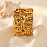 A wide gold-plated cuff bracelet with detailed religious engravings, displayed on a cream satin fabric with a seashell prop in the background.