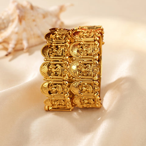 A wide gold-plated cuff bracelet with detailed religious engravings, displayed on a cream satin fabric with a seashell prop in the background.