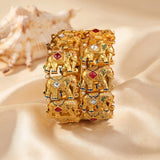 A wide gold-toned bangle bracelet featuring a repeating relief pattern of decorated elephants adorned with red, green, and white stone-like accents, displayed on draped cream satin fabric with a seashell prop in the background.