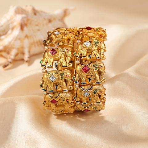 A wide gold-toned bangle bracelet featuring a repeating relief pattern of decorated elephants adorned with red, green, and white stone-like accents, displayed on draped cream satin fabric with a seashell prop in the background.